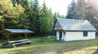 Blick auf eine kleine massiv gebaute Waldhütte mit überdachter Sitzgruppe davor. im Hintergrund ist Wald.