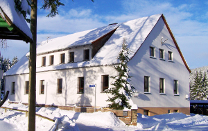 Forstamt im Stil eines Wohnhauses. Es ist Winter und das Haus und die Straße und der Gehweg sind mit Schnee bedeckt. Im Hintergrund verschneite Nadelbäume.